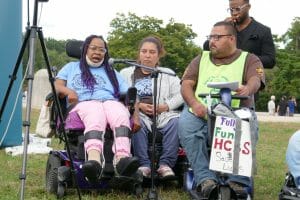 Three people in wheelchairs speak in front of a microphone holding a sign that "Fully fund home-based 