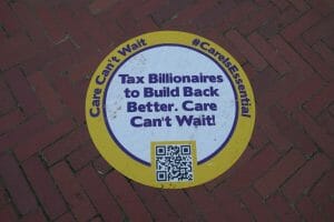 A circular placard on the sidewalk reads "Tax Billionaires to Build Back Better, Care Can't Wait!" in blue text.
